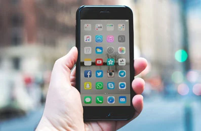 Hand holding black iPhone displaying colorful home screen with social media and messaging apps on blurred city street background